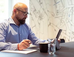Man with glasses and a beard working intently on a tablet at a desk with a notepad, coffee cup, and glass of water.